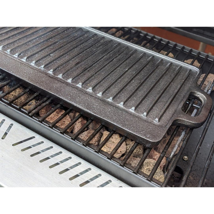 A close-up of the Black Rock Grill Large Reversible Cast Iron Griddle Pan with silicone grip handles (46cm x 23cm) on a gas grill grate, with lava rocks beneath, plus marinating oil brush included.