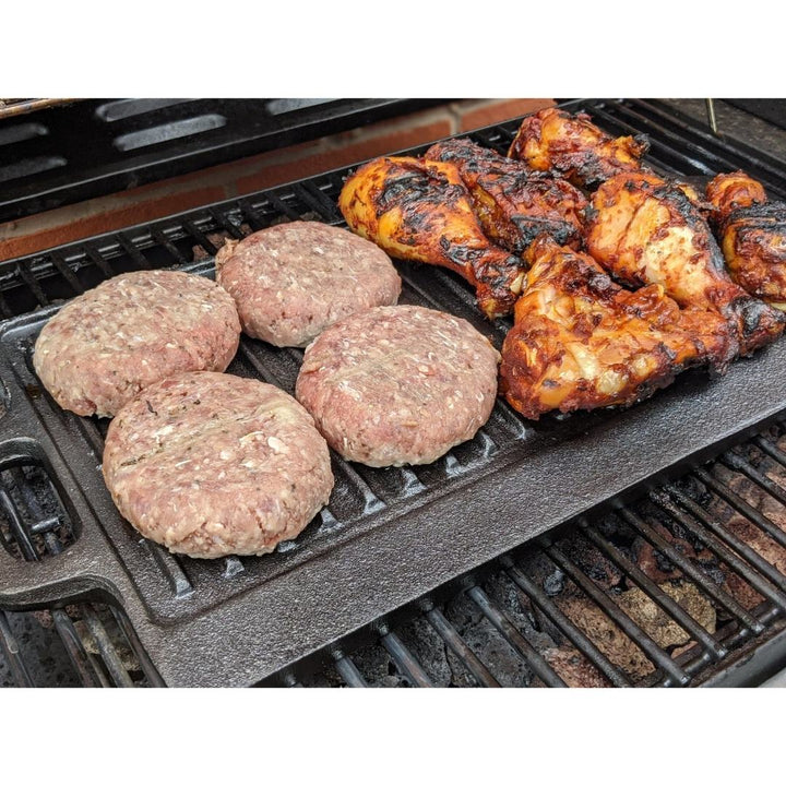 Raw burger patties and charred chicken cook side by side on the Black Rock Grill Large Reversible Cast Iron Griddle Pan (46cm x 23cm) with silicone grip handles, locking in flavor and giving perfect grill marks. Includes marinating oil brush.