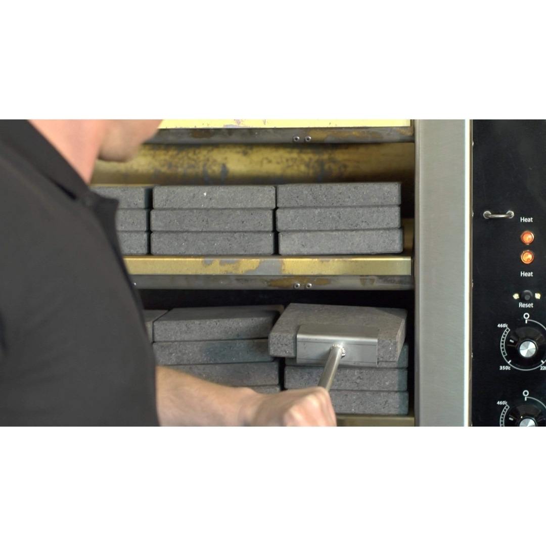 A person carefully adjusts the Black Rock Grills Rock Shovel for Steak Stones in a furnace with insulated bricks, using a long handle for precision. Control knobs and indicators are visible on the right side to ensure perfect settings.