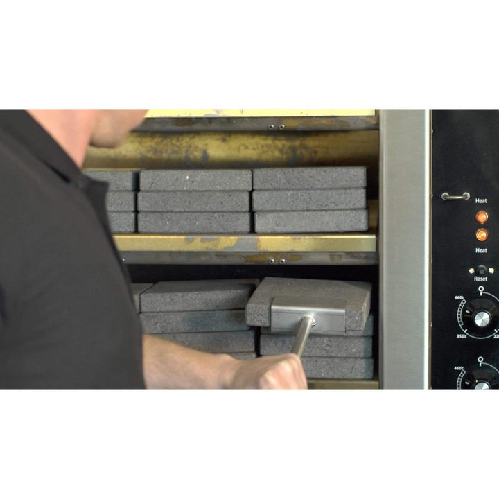 A person carefully adjusts the Black Rock Grills Rock Shovel for Steak Stones in a furnace with insulated bricks, using a long handle for precision. Control knobs and indicators are visible on the right side to ensure perfect settings.