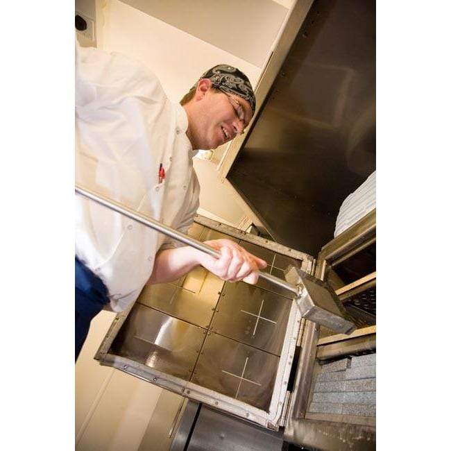 A chef in a white uniform and bandana uses a Black Rock Grill Rock Shovel for Steak Stones to open a large industrial oven, revealing stacks of plates inside.