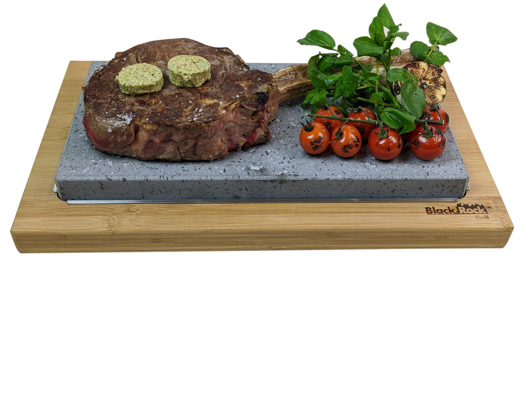 A sizzling bone-in steak with herb butter sits on trendy lava stones, complemented by vine tomatoes and greens. This unique hot stone cooking experience is elegantly presented on a wooden board featuring the Black Rock Grill Steak Stone Set for 2.