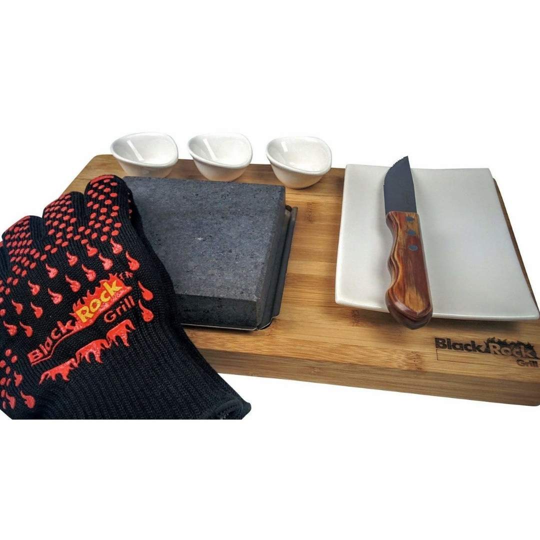The Black Rock Grill Steak On The Stone Set lets you cook your own meal and includes a black grill glove, large stone slab, wooden knife with a ceramic blade, rectangular white plate, and three small white sauce dishes on a Black Rock Grill marked wooden board.