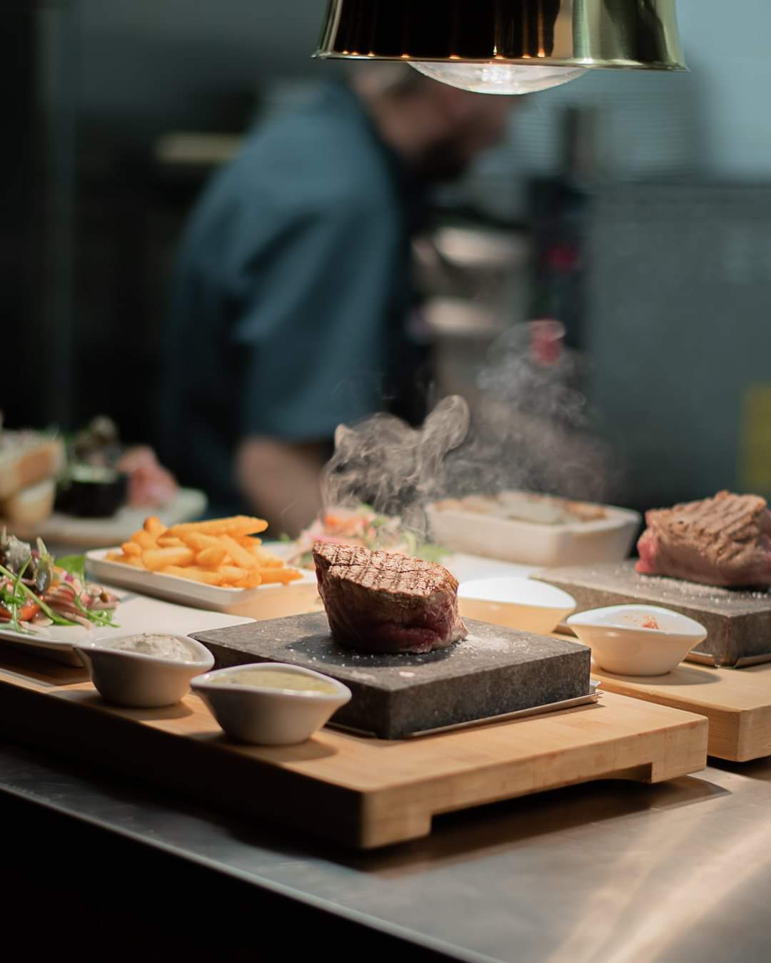 The Black Rock Grills ROXY12 set serves a sizzling steak on a hot stone for an interactive meal, accompanied by fries, vegetables, and sauces. Warm overhead lighting and a blurred kitchen figure add to the ambiance.
