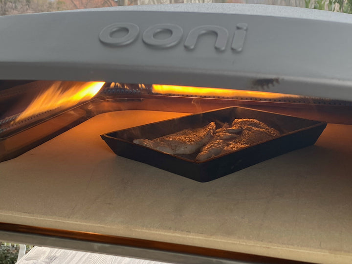 A visible flame cooks seasoned fish on a sizzling Cast Iron Fajita Skillet Pan with Wood Base from Black Rock Grill in an open pizza oven, showing ooni on top, with the tray resting on a stone surface inside.