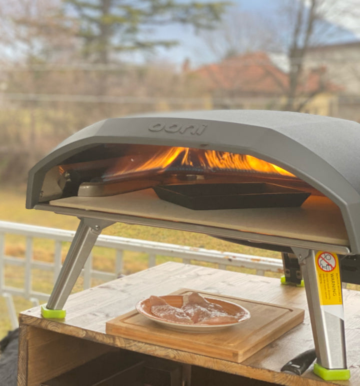 A premium Black Rock Grill outdoor pizza oven with a visible flame is set up on a wooden stand. It features a pizza stone inside, while the Cast Iron Fajita Skillet Pan with Wood Base sits below, displaying raw meat wrapped in plastic. Trees and a fence are visible in the background.