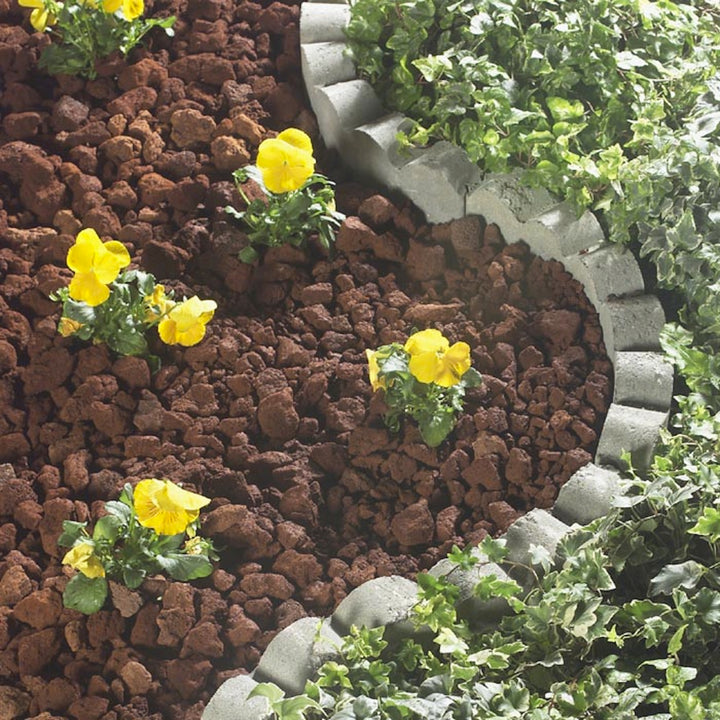 Yellow flowers in a garden bed are encircled by brown mulch, bordered by gray stones and green ivy. The semi-circular, horticulture-inspired design is enhanced with Black Rock Grills Large Lava Rock (8-15cm), creating a neat and vibrant landscape.