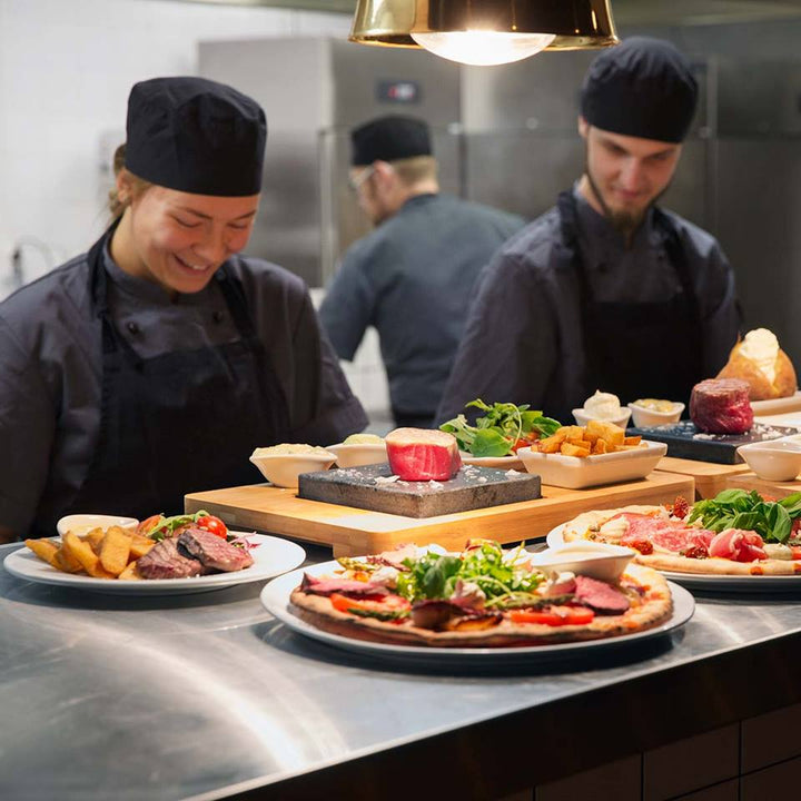 In a vibrant restaurant kitchen, chefs skillfully prepare dishes using the Black Rock Grill ROXY12 set. A female chef smiles while two male chefs focus on healthy dining, with plates of pizza and steak garnished with fresh ingredients under warm lighting.