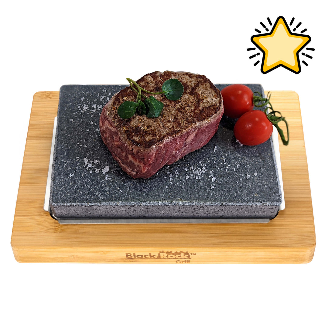 A cooked steak garnished with herbs and coarse salt sits on a black stone slab with two vine tomatoes, all on a wooden board labeled Black Rock Grill. A yellow star icon is in the top right corner.