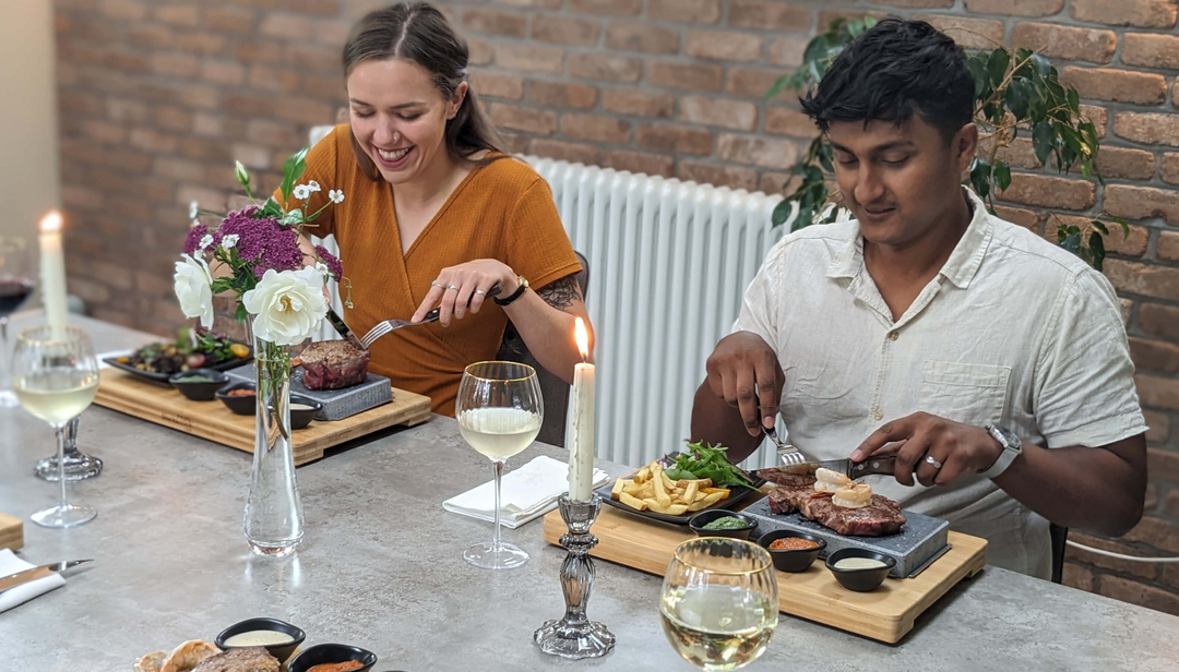 Two people sit at a table with wine and steak dinners, smiling and enjoying their meal. The table is set with flowers, candles, and various side dishes. A brick wall and plants are in the background.