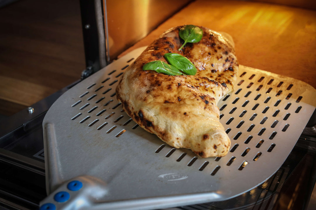 A freshly baked calzone with golden spots and basil sits on a metal peel, being taken out of the Macte Voyager Twin Electric Pizza Oven—an AVPN Certified Italian countertop oven by Macte.