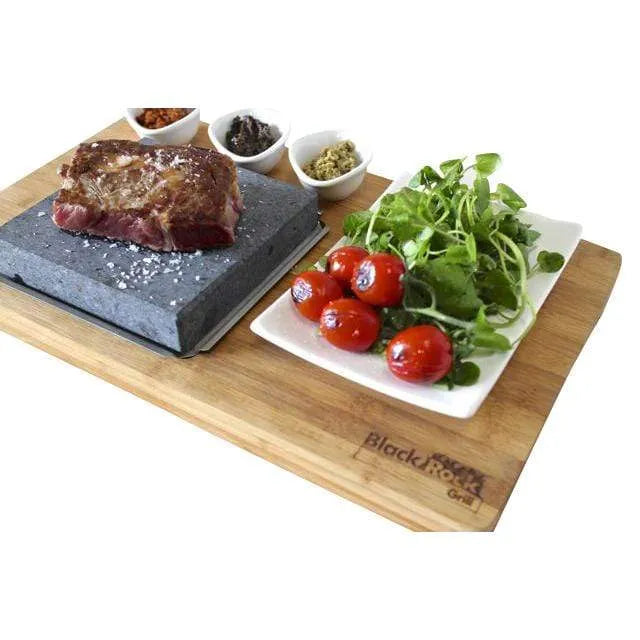 Steak Stone on underplate and wooden bamboo board. On the steak stone is a steak sprinkled with salt. The Bamboo Board also has 3 Matt Black sauce bowls and Matt Black side plate containing salad and tomatoes