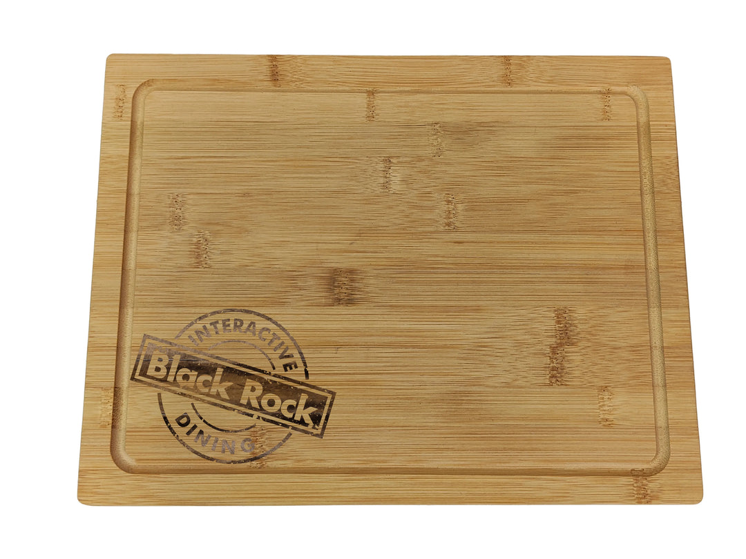 Bamboo cutting board with the words Black Rock Interactive Dining stamped on the lower left corner. The board has a smooth, light wood grain and a shallow groove near the edge.