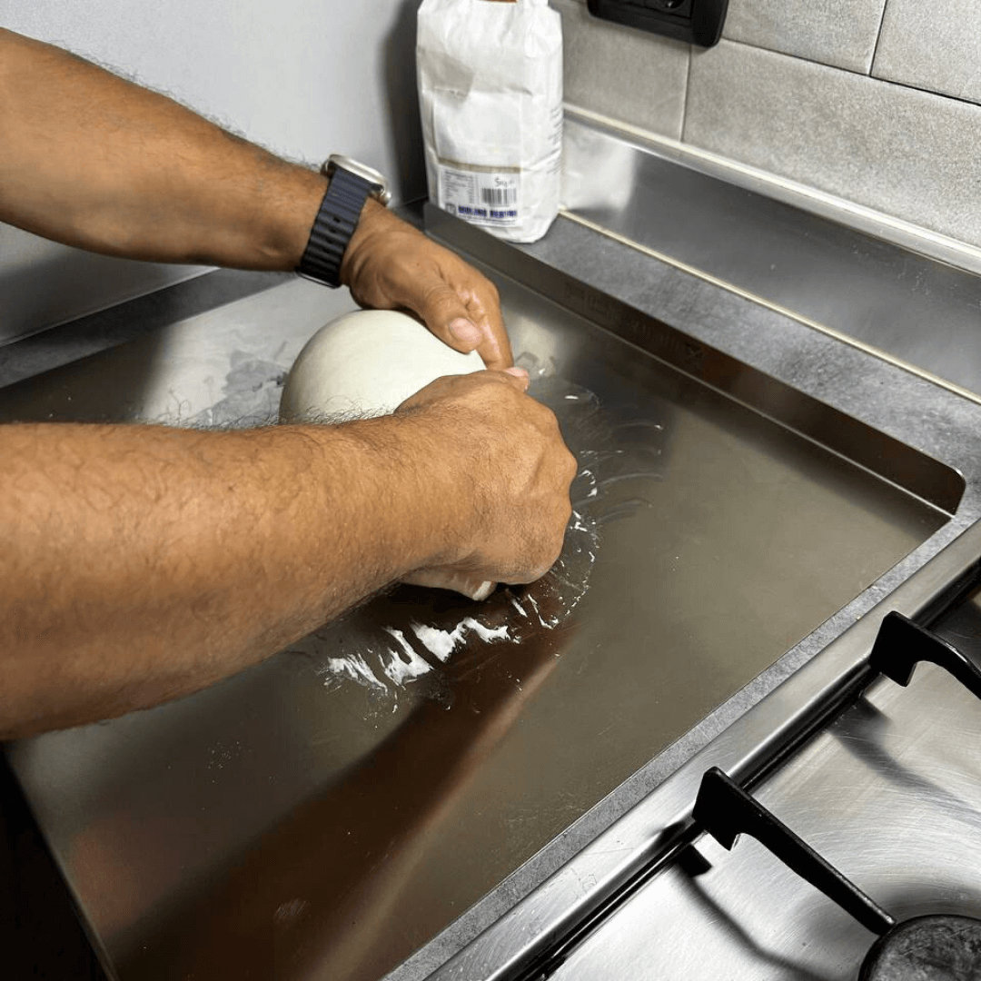 In the kitchen, someone expertly kneads dough on the Effeuno Stainless Steel Pizza Prep Board, Professional Food-Grade AISI 304, 50cm, surrounded by scattered flour. Nearby, a stove sits idle and a package of flour is seen in the background. The person wears a ticking watch on their left wrist.