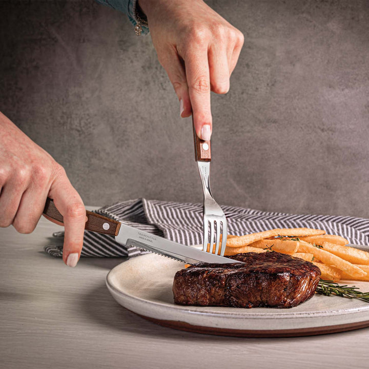 Hands holding a fork and knife from the Tramontina 12 Piece Steak Cutlery Set are slicing a cooked steak on a plate, accompanied by golden-brown fries. A striped cloth napkin appears in the background on a gray surface.