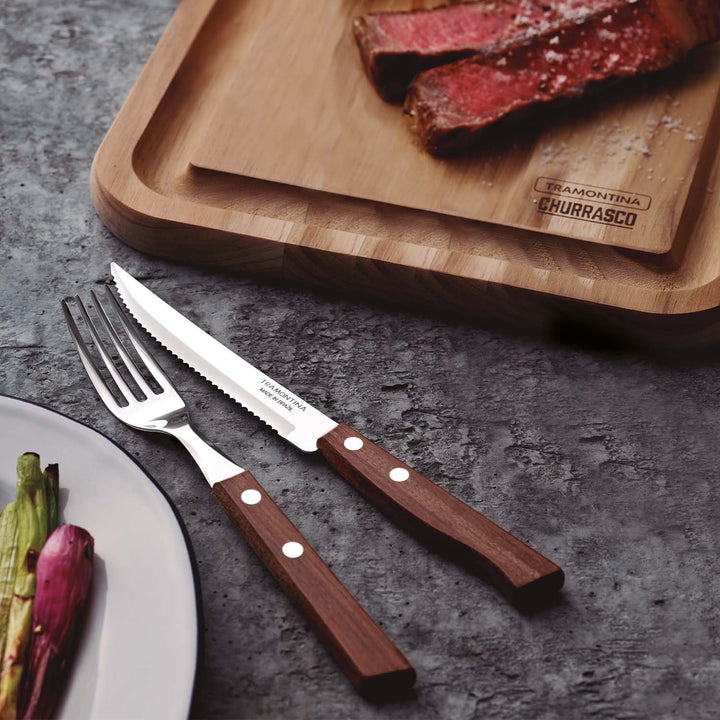 In the background, sliced steak sits on a wooden cutting board. In the foreground, a stainless steel fork and serrated knife from the Tramontina 12 Piece Steak Cutlery Set with polywood handles lie next to a plate of grilled vegetables.