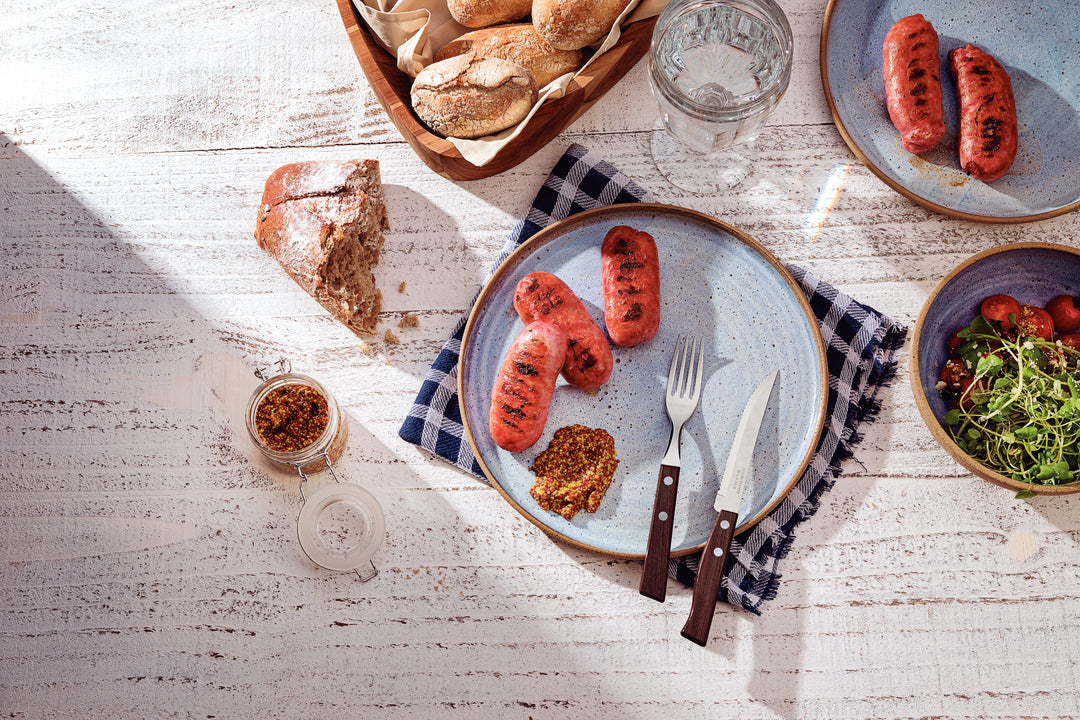 A tabletop features grilled sausages on a plate with mustard, using a sleek Tramontina stainless steel knife and fork. A basket of bread rolls, a glass of water, a bread slice, and a salad bowl complete the scene atop the checkered cloth that adds rustic charm.