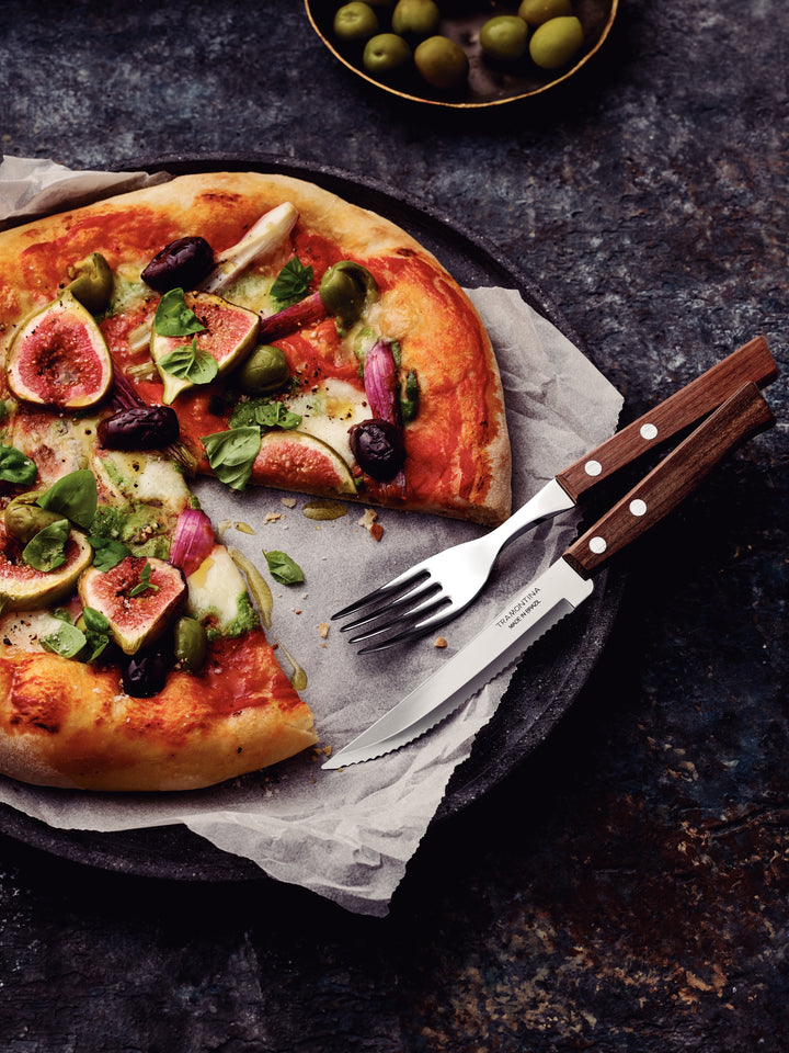 A freshly baked pizza with figs, olives, basil, and cheese is on a parchment-lined plate. Nearby, Tramontina 12 Piece Steak Cutlery Set-Service for 6 rests at the side. A small bowl of green olives appears in the background on a dark textured surface.