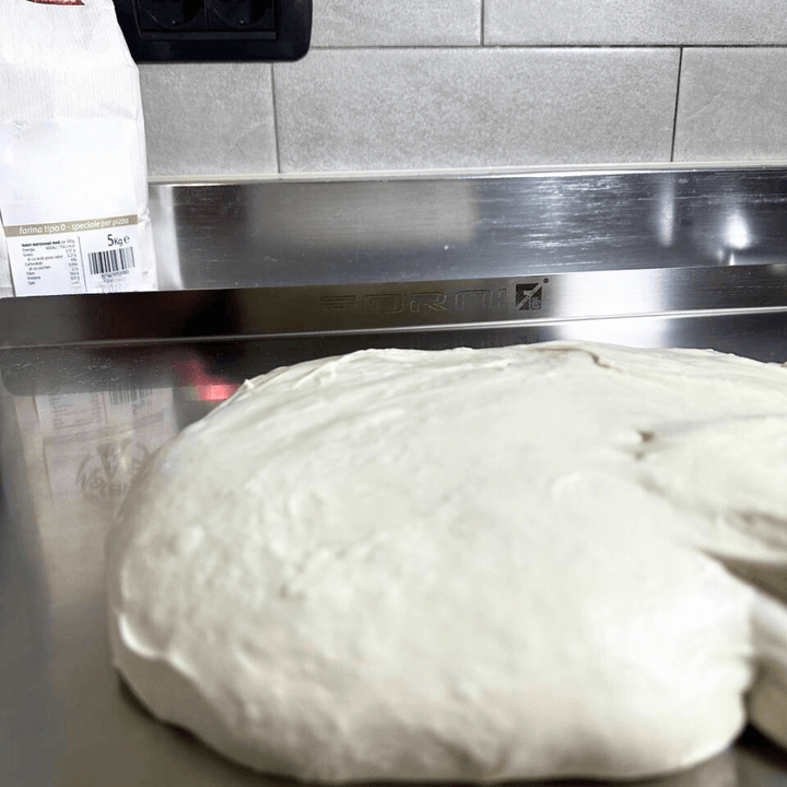 A ball of pale dough rests on the Effeuno Stainless Steel Pizza Prep Board – Professional Food-Grade AISI 304, 50cm, in the kitchen. A bag of flour is partially visible against gray tiled walls, creating an ideal setup for using this top-grade pizza-making tool.