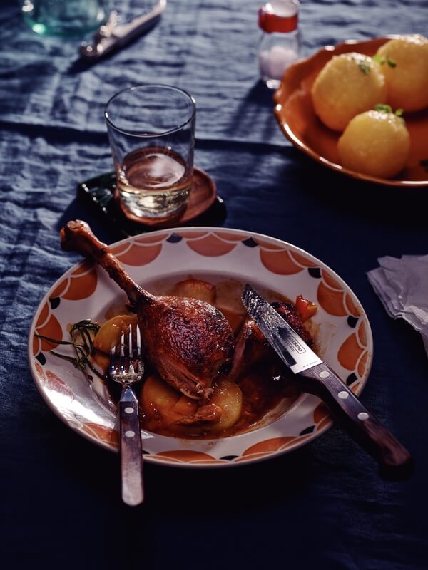 A plate with roasted duck leg, potatoes, and sauce sits on a blue tablecloth beside Black Rock Grill's Tramontina Gaucho Premium Brazilian Steak Knives, a glass of water, cutlery, and a bowl of round dumplings.