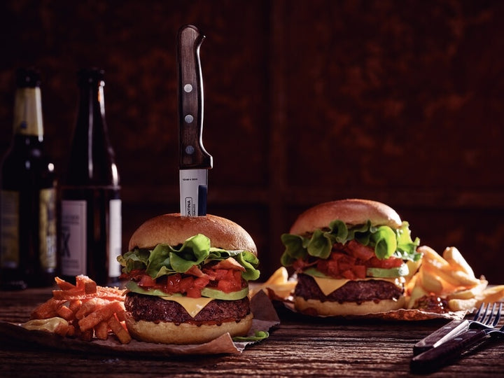 Two cheeseburgers with lettuce, cheese, and tomato on buns sit on a wooden table with fries; a Tramontina Gaucho Premium Brazilian Steak Knife by Black Rock Grill is stabbed into one burger. Beer bottles and utensils are in the background.