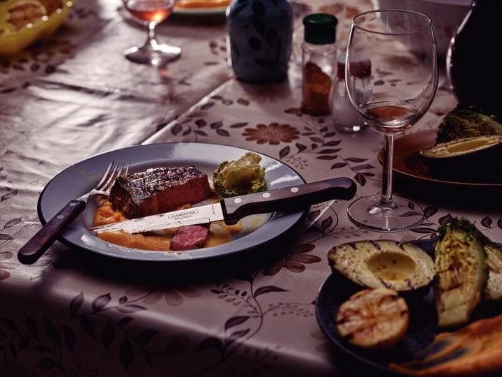 A floral-patterned tablecloth holds a plate with steak, mashed potatoes, and vegetables. Tramontina Gaucho Premium Brazilian Steak Knives by Black Rock Grill rest on the plate, alongside a wine glass, grilled avocado, and condiments.