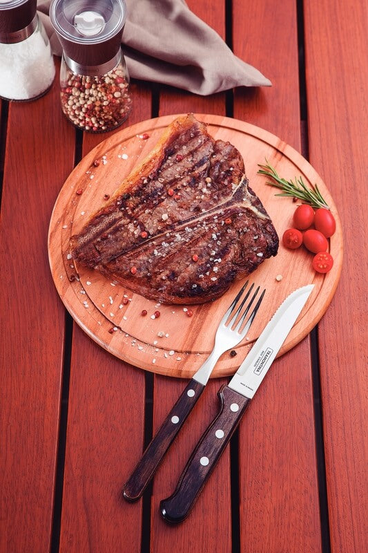 A juicy grilled steak on a wooden board, garnished with salt, peppercorns, cherry tomatoes, and rosemary, sits alongside a fork and Black Rock Grill's Tramontina Gaucho Premium Brazilian Steak Knives on a red wooden table.