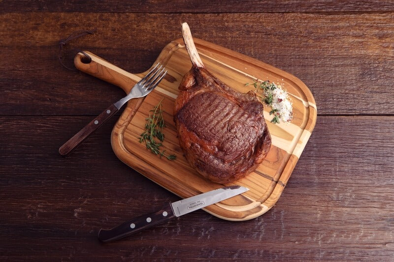 A cooked tomahawk steak sits on a wooden board with herbs, coarse salt, and pepper. Black Rock Grill Tramontina Gaucho Premium Brazilian Steak Knives and cutlery are placed beside it on a rustic wooden table.