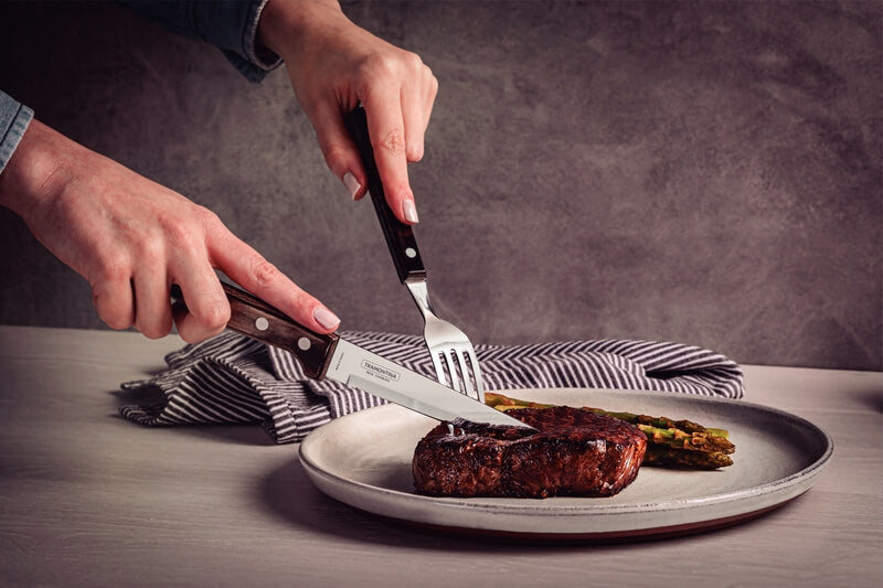 A person uses Black Rock Grill Tramontina Gaucho Premium Brazilian Steak Knives to cut a cooked steak with asparagus on a plate, on a table with a striped napkin in the background.