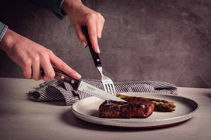 A person uses Black Rock Grill Tramontina Gaucho Premium Brazilian Steak Knives to cut a cooked steak with asparagus on a plate, on a table with a striped napkin in the background.