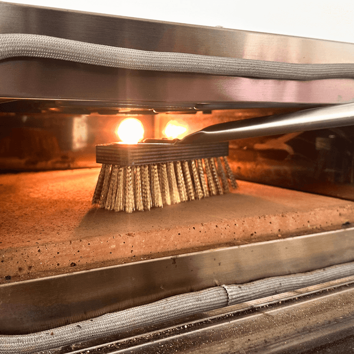 The Effeuno Pizza Oven Brush, featuring a long handle and brass bristles, efficiently cleans by contacting the stone surface, while two bright lights illuminate and reflect off interior metal surfaces.