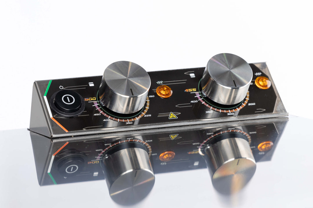 A close-up of the Macte Voyager Twin Electric Pizza Oven's control panel shows two large metal knobs, colored indicator lights, a power switch, and measurement markings reflected on a shiny surface. AVPN Certified Italian Countertop Oven by Macte.