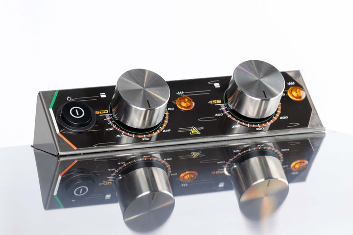 A close-up of the Macte Voyager Twin Electric Pizza Oven's control panel shows two large metal knobs, colored indicator lights, a power switch, and measurement markings reflected on a shiny surface. AVPN Certified Italian Countertop Oven by Macte.