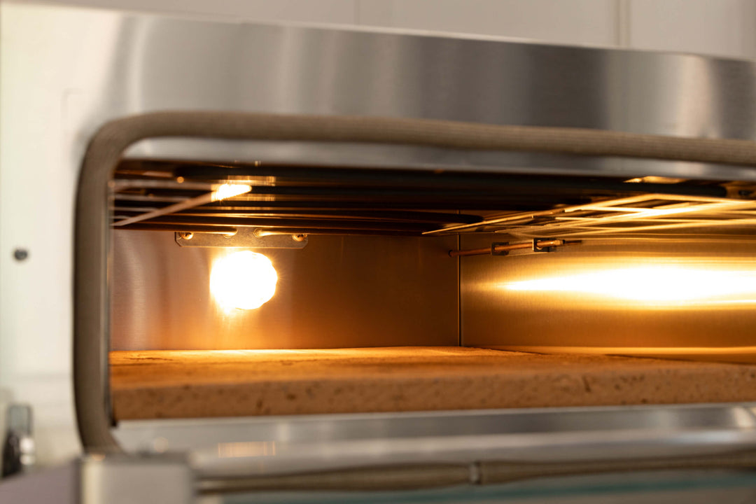 A close-up of the Macte Voyager Twin Electric Pizza Oven by Macte, an AVPN Certified Italian countertop oven, with its interior light on, showing empty metal racks and a stone baking surface.