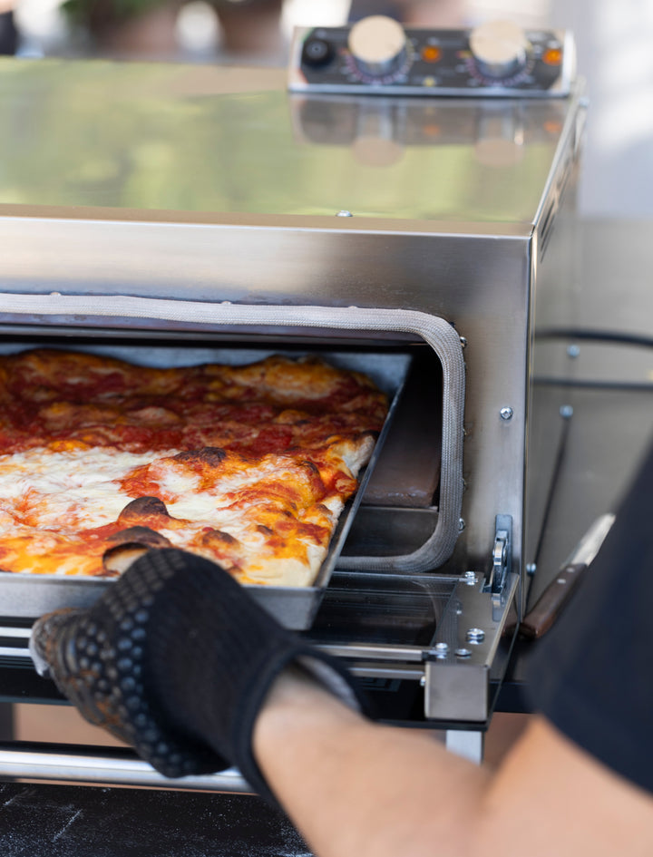 Wearing a black oven mitt, a person removes a rectangular pizza with melted cheese and sauce from the Macte Nettuno Electric Pizza Oven, an authentic Italian countertop oven for pizza and bread by Macte.