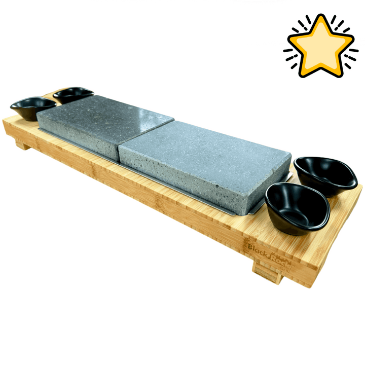 The Black Rock Grill Signature Steak Sharing Stone Set features a rectangular bamboo platter holding two large gray steak stones in the center and four small black dipping bowls at each corner, with a yellow hand-drawn star graphic in the upper right.