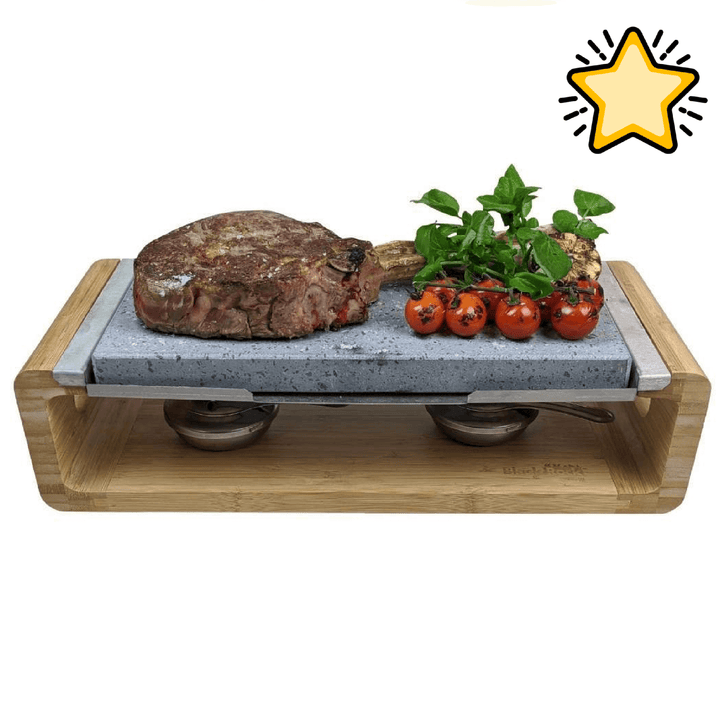 A cooked steak and grilled cherry tomatoes with greens are served on the Black Rock Grill Steak Stone Sharing Platter & Grill Set, which sits on a wooden tray with metal burners. A large yellow star icon appears in the top right corner.