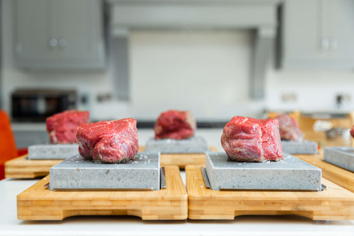 Black Rock Grill’s Sizzling Steak Stone 20 Rock Set Summer Matchday Kit showcases raw beef steaks on hot stones atop wooden trays, creating an inviting dining experience on a modern kitchen countertop with grey cabinets.