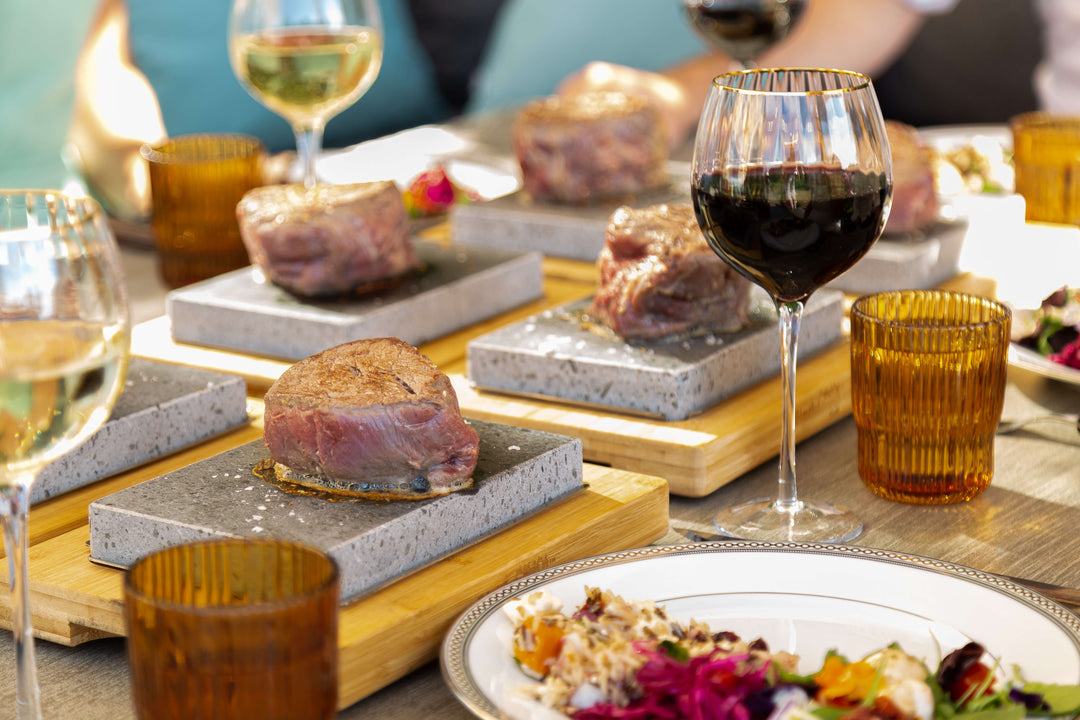 The Black Rock Grill Sizzling Steak Stone 20 Rock Set Summer Matchday Kit presents cooked steaks on stone slabs, paired with amber glasses, red and white wine, and a salad plate in the foreground on the dining table.