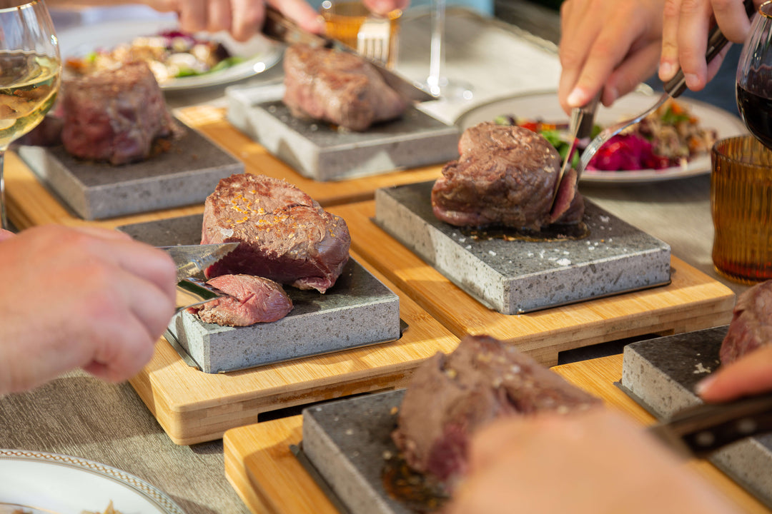 Several people use the Black Rock Grill Sizzling Steak Stone 20 Rock Set Summer Matchday Kit to cut and enjoy cooked steaks served on hot stones atop wooden boards, creating an interactive dining experience with wine and salad in the background.