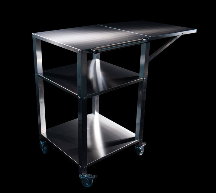 The Effeuno Pizza Oven Stand & Prep Table – CR55 is a stainless steel work trolley with an adjustable shelf, ideal as a mobile workstation. It features four wheels for easy mobility and has a sleek metallic finish that contrasts against its black backdrop.