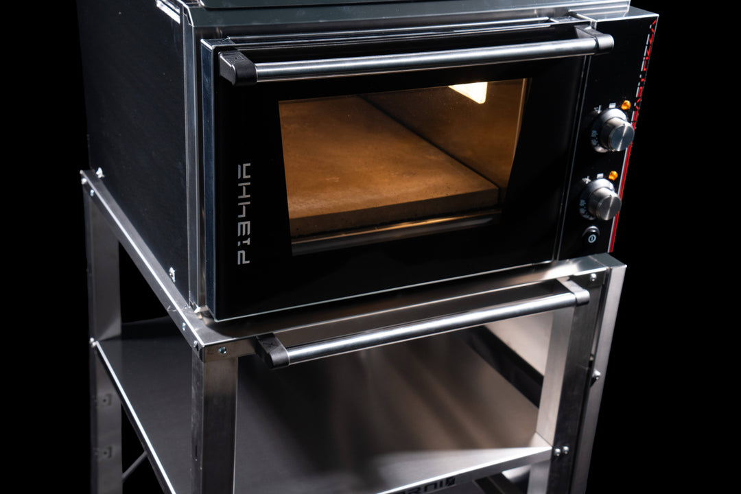 The Effeuno Pizza Oven Stand & Prep Table features a black and silver oven mounted on the CR55 Stainless Steel Work Trolley with an adjustable shelf. The oven has a transparent door, two right-side dials, and an illuminated interior.