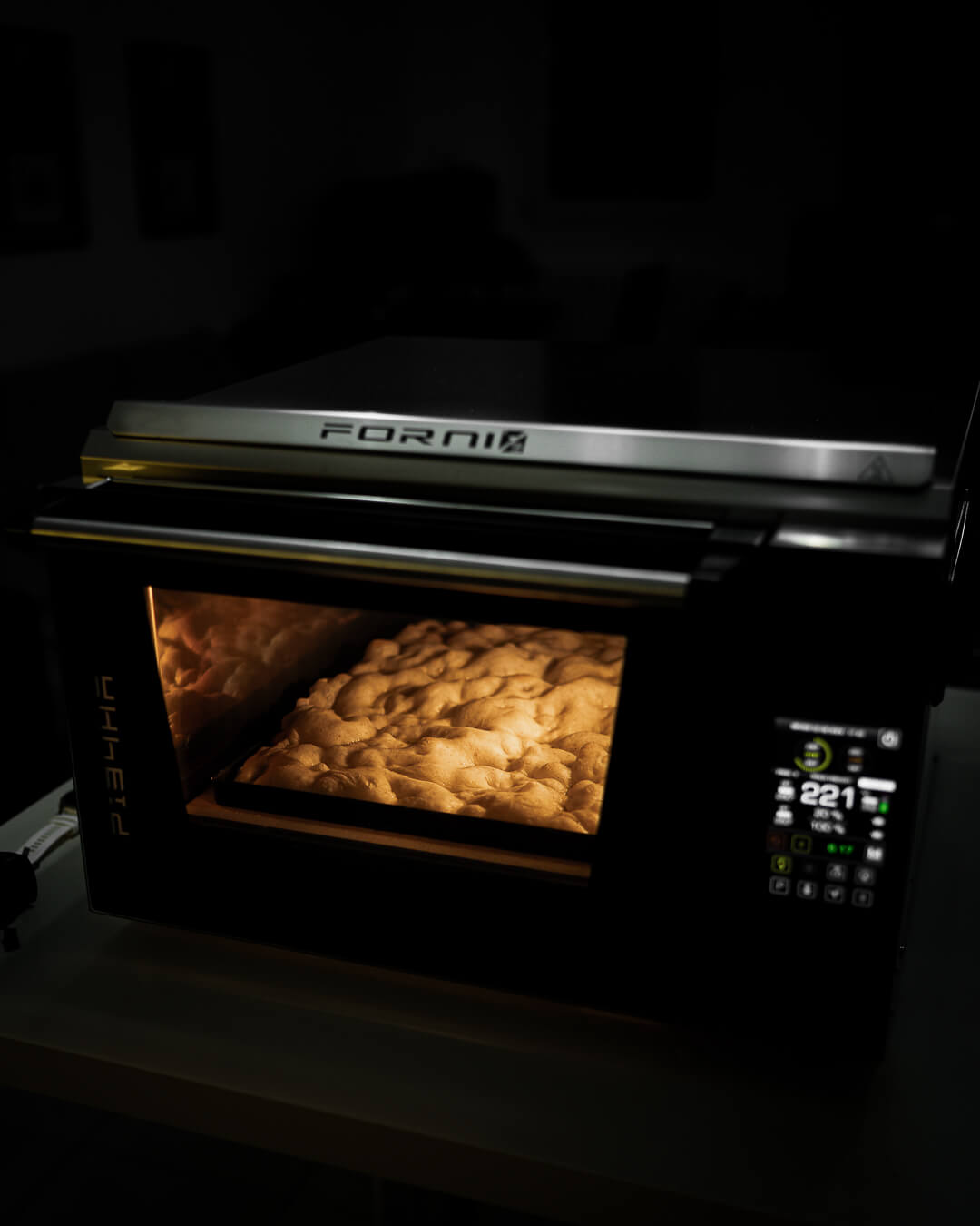 A modern black Forni oven, handcrafted in Italy, bakes bread to perfection with a warm glow highlighting loaves beside an Effeuno Professional Rectangle Blue Iron Pizza Pan. The digital display shows 221 degrees, as the oven sits on a countertop in a dimly lit room.