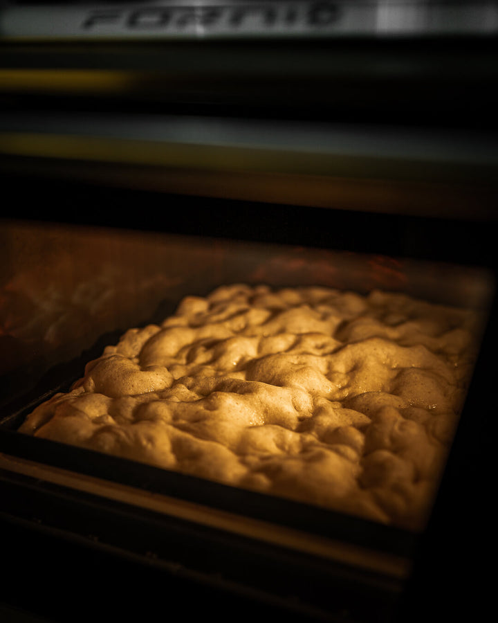 Golden brown focaccia bread bakes, its textured surface glowing warmly. Crafted with care as if from Italy, it rests on an Effeuno Professional Rectangle Blue Iron Pizza Pan with Diagonal X Antfelexion 30x40 x2cm, nodding to traditional Roman and focaccia pizza styles.