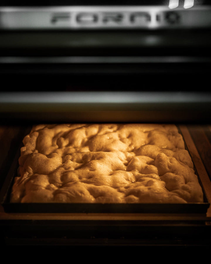 A golden brown focaccia, reminiscent of Roman and focaccia pizza, bakes on the Effeuno Professional Rectangle Blue Iron Pizza Pan with Diagonal X Antfelexion. Its dimpled surface glows under the oven light, adding a touch of Italy to your kitchen as the oven door peeks from above.