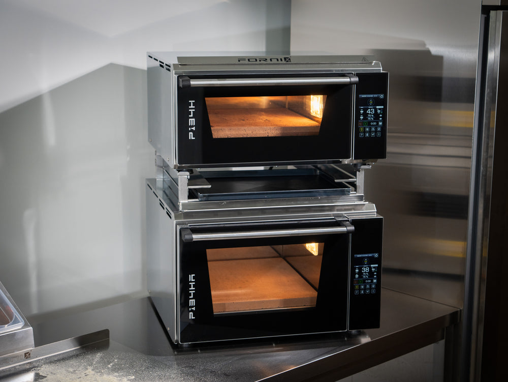 Two Effeuno PRO & Evolution ovens are stacked on a stainless steel table using the Black Rock Grill Stacking Kit for a safe double oven mount. Their digital panels are lit, showing empty stone baking surfaces inside.