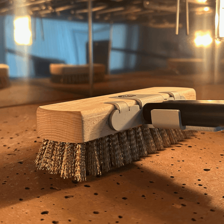 A close-up of an effeuno pizza oven brush head with a black handle and brass bristles, shown on a biscotto stone textured surface in pizza oven with blurred lights in the background.
