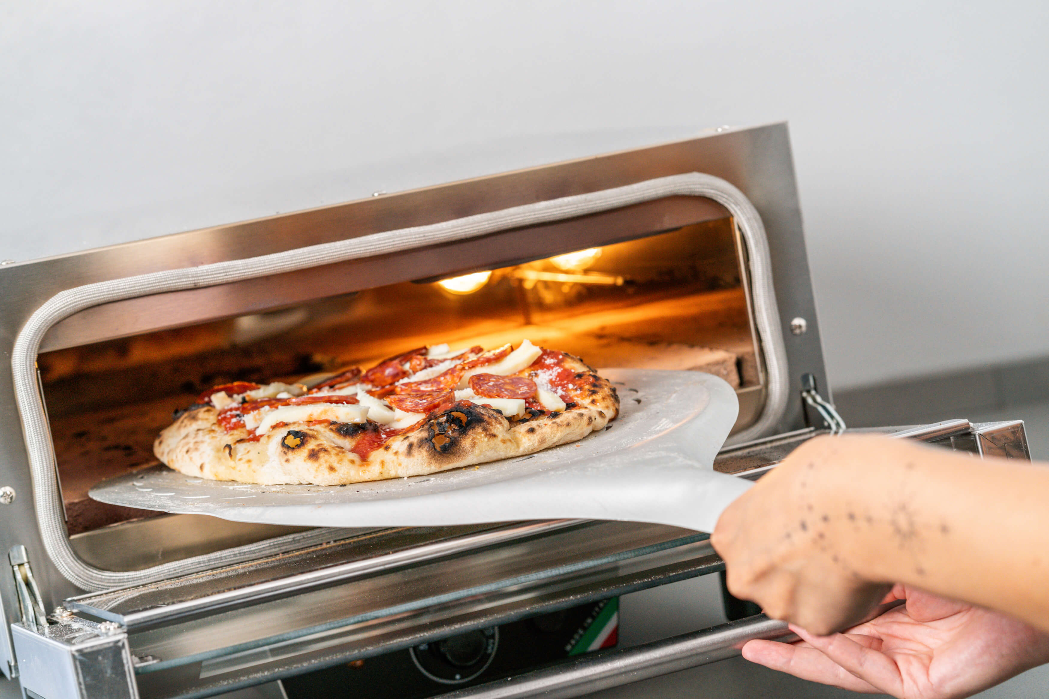 A hand uses a metal pizza peel to remove a cheese and pepperoni Neapolitan pizza from the Effeuno N3 Electric Pizza Oven with biscotto stone, made in Italy, as the oven’s light highlights the hot crust and toppings.