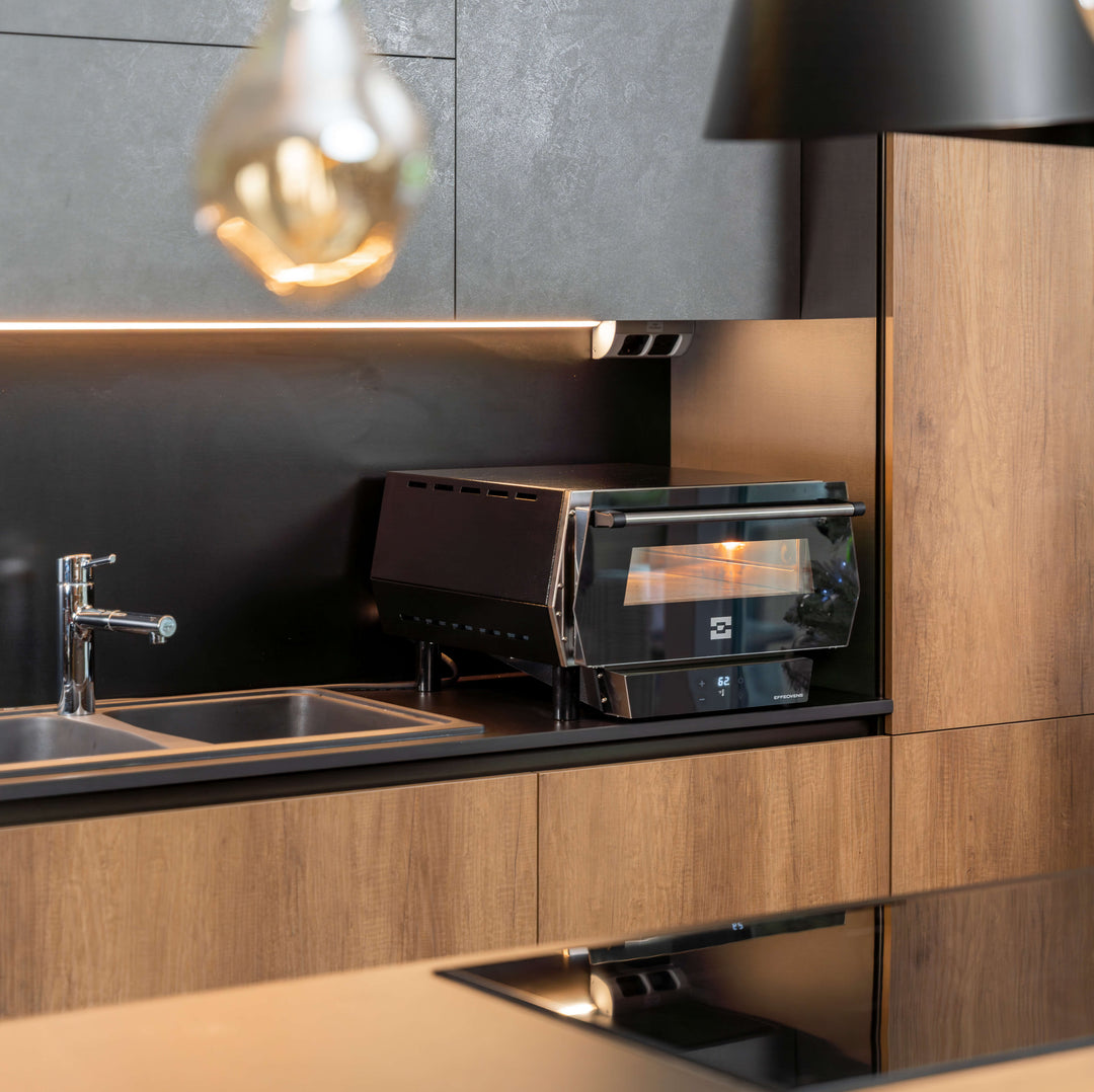 A modern kitchen features an Effeuno Pulse Electric Pizza Oven, UL listed and built-in, next to a stainless steel sink with faucet, set against wood and black cabinets with under-cabinet lighting.