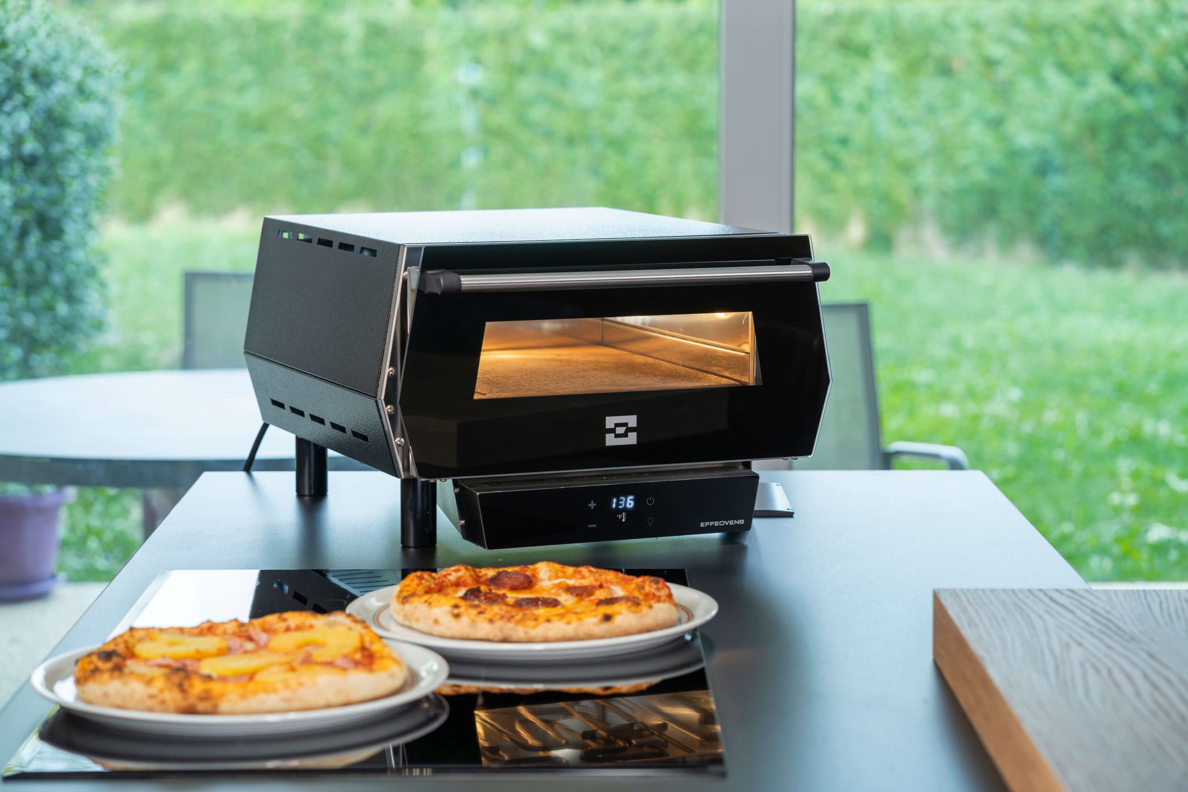 An Effeuno Pulse Electric Pizza Oven – 948F / 509°C with Biscotto Stone, made in Italy, stands on a kitchen island with two pizzas in front. Large windows behind it reveal a lush outdoor scene. Suitable for indoor or outdoor use.